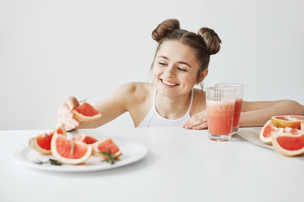 Bella donna felice che sorride prendendo fetta di pompelmo dal piatto che si siede alla tavola sopra la parete bianca. Cibo fitness sano.