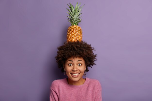 Bella donna felice con taglio di capelli afro, tiene ananas fresco con foglie verdi sulla testa, posa con frutta estiva