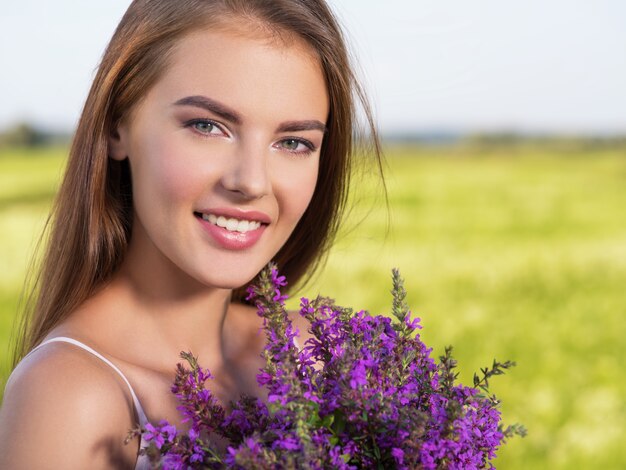 Bella donna felice e sorridente all'aperto con fiori viola nelle mani. La giovane ragazza allegra è sulla natura sopra il campo di primavera. Concetto di libertà. Ritratto di un modello carino e sexy sul prato