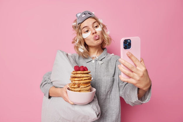 Bella donna imbronciata labbra e pose alla fotocamera frontale dello smartphone posa con deliziose frittelle per colazione indossa un pigiama tiene morbido cuscino applica cerotti di bellezza pone contro il muro rosa