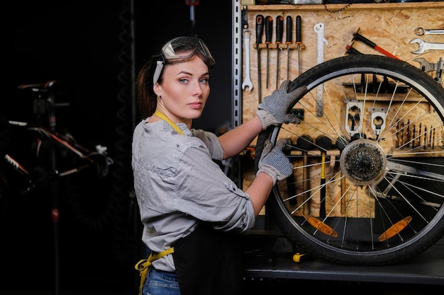 Bella donna in abiti da lavoro, grembiule e occhiali, ripara una ruota di bicicletta in un'officina.