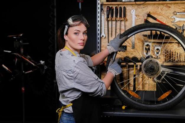 Bella donna in abiti da lavoro, grembiule e occhiali, ripara una ruota di bicicletta in un'officina.