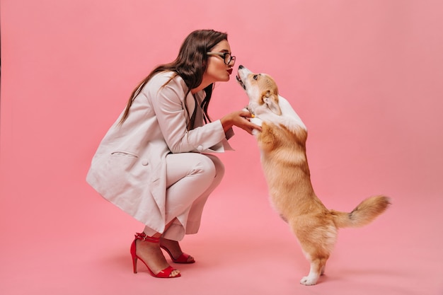 Bella donna in abito beige gioca con il cane su sfondo rosa. Signora carina di affari in vestito elegante e scarpe rosse bacia corgi.