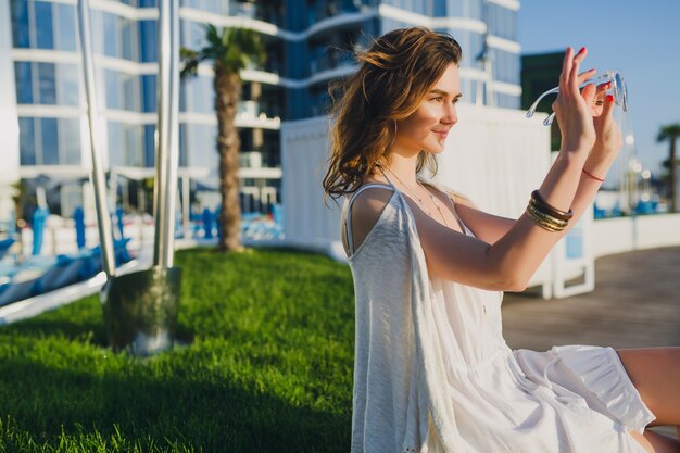 Bella donna in abito bianco in hotel resort estivo, indossando occhiali da sole e accessori alla moda, ridendo, vacanze