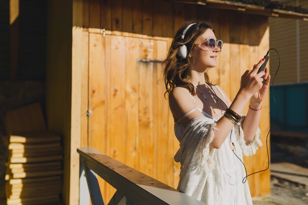 Bella donna in abito estivo bianco ascoltando musica in cuffia ballando e divertendosi, tenendo lo smartphone, stile di vacanze estive
