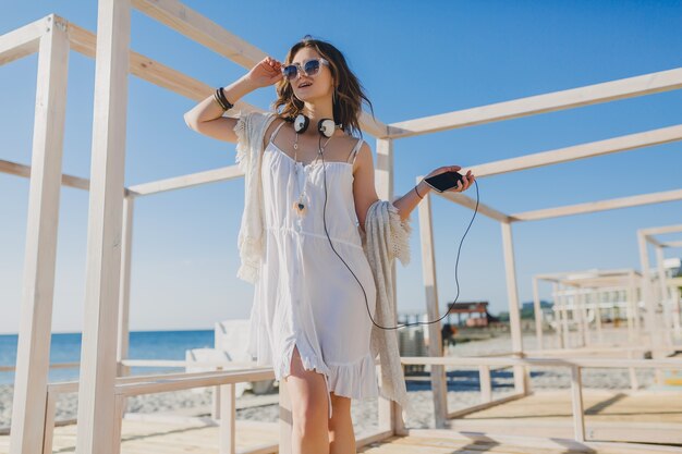Bella donna in abito estivo bianco ascoltando musica in cuffia ballando e divertendosi, tenendo smartphone, spiaggia di mare stile vacanze estive