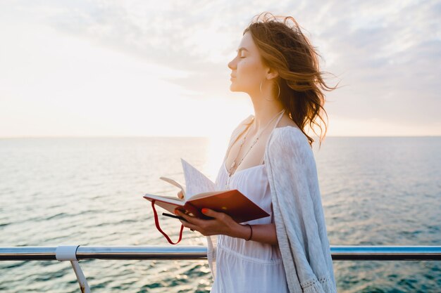 Bella donna in abito estivo bianco che cammina in riva al mare all'alba con il libro del diario in stato d'animo romantico pensando e prendendo appunti