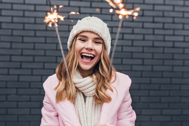 Bella donna in accessori invernali che esprimono emozioni positive e agitando le stelle filanti. Foto all'aperto della meravigliosa ragazza con cappello rosa che celebra il nuovo anno.