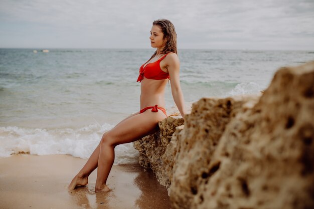 Bella donna in bikini rosso che posa sulla spiaggia che si siede sulle rocce