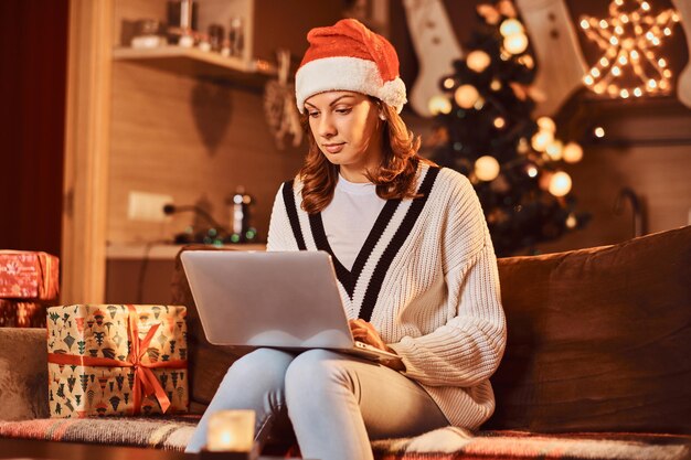 Bella donna in cappello che si rilassa sul divano facendo acquisti di Natale su internet nella stanza decorata nel periodo natalizio.