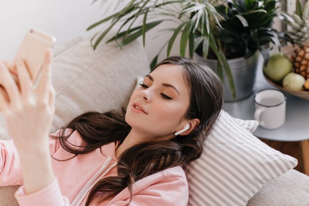Bella donna in cuffie wireless ascoltando musica, sdraiato sul divano e chiacchierando in smartphone