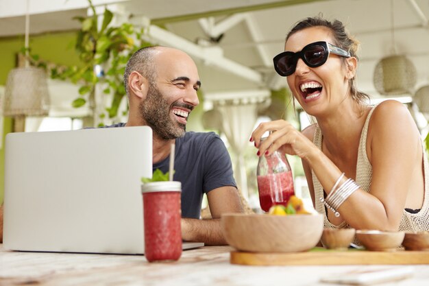 Bella donna in occhiali da sole alla moda che tiene un bicchiere di succo di frutta fresco e ride mentre il suo bel amico con la barba le racconta una storia divertente.