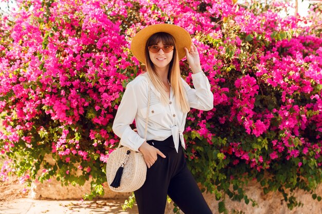 Bella donna in piedi sui fiori rosa Indossando cappello di paglia e abbigliamento casual.