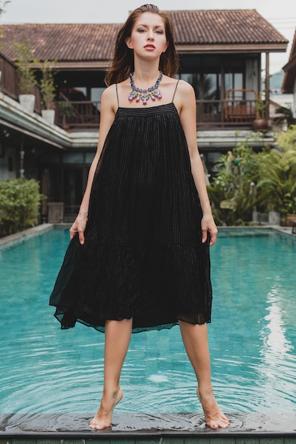 Bella donna in vestito nero lungo alla moda che propone alla piscina della villa tropicale, stile estivo elegante, vacanze, tendenza della moda, camminare a piedi nudi