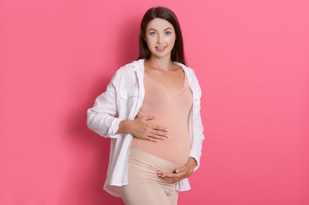 Bella donna incinta che abbraccia la sua pancia e che guarda l'obbiettivo con calma espressione facciale felice, signora con un aspetto piacevole che indossa abiti casual tocca la sua pancia.