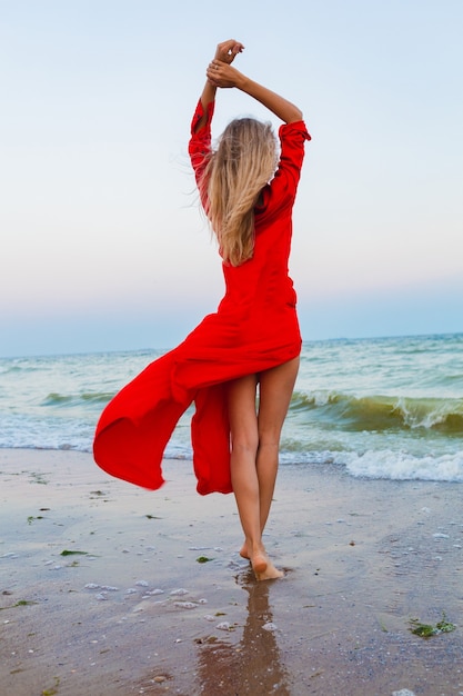 Bella donna libera in vestito rosso nel vento sulla spiaggia del mare che cammina in estate