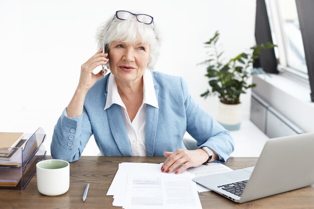 Bella donna matura con capelli grigi che fa telefonate nel suo ufficio, imprenditore femminile senior elegante in vestito alla moda parlando sul cellulare al potenziale partner, seduto al posto di lavoro con il computer portatile