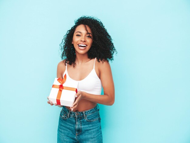 Bella donna nera con l'acconciatura di riccioli afro Modello sorridente vestito con abiti estivi bianchi Femmina spensierata sexy in posa vicino al muro blu in studio Confezione regalo abbronzata e allegra Holding isolata