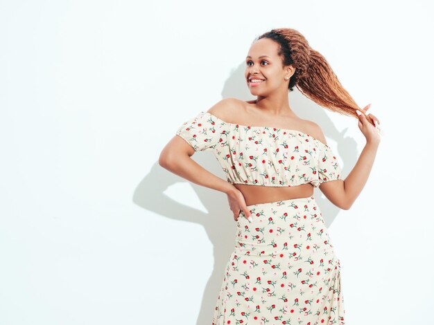 Bella donna nera con l'acconciatura di riccioli afro Modello sorridente vestito in abito estivo bianco Femmina spensierata sexy in posa vicino al muro in studio Abbronzato e allegro Isolato