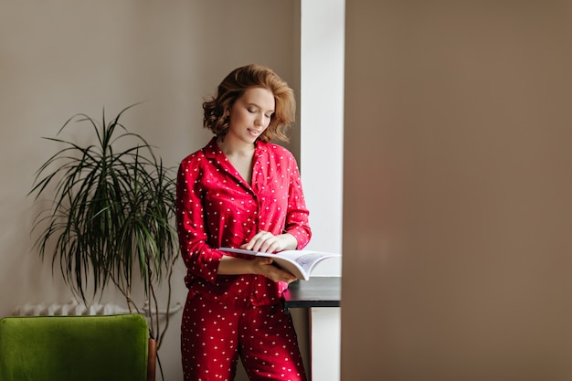 Bella donna pensierosa che legge la rivista di mattina. Tiro al coperto di donna attraente in indumenti da notte rossi.