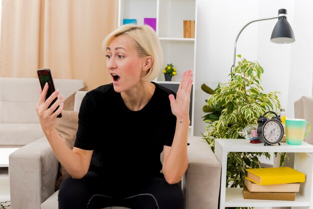 Bella donna russa bionda scioccata si siede sulla poltrona alzando la mano e guardando il telefono all'interno del soggiorno