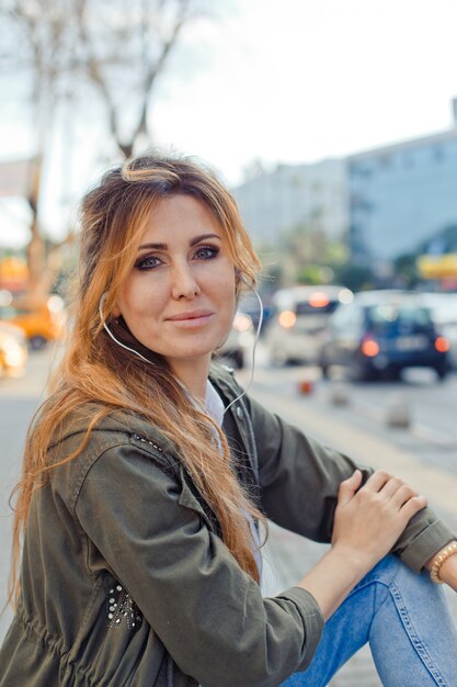 Bella donna seduta su una panchina leggermente sorridente e ascolto musica in strada durante il giorno
