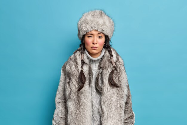 Bella donna seria concentrata sul davanti indossa abiti cappotto cappello invernale per pose fredde sul muro blu. Ragazza di Natale in capispalla