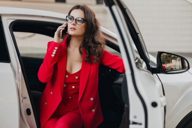Bella donna sexy ricca di affari in vestito rosso che si siede in macchina bianca, con gli occhiali parlando al telefono, stile donna d'affari