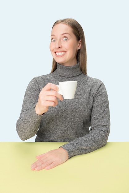 Bella donna sola che si siede allo studio blu e che sembra triste tenendo in mano la tazza di caffè. Closeup ritratto tonico in stile minimalista