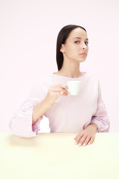 Bella donna sola che si siede allo studio rosa e che sembra triste tenendo in mano la tazza di caffè. Closeup ritratto tonico in stile minimalista