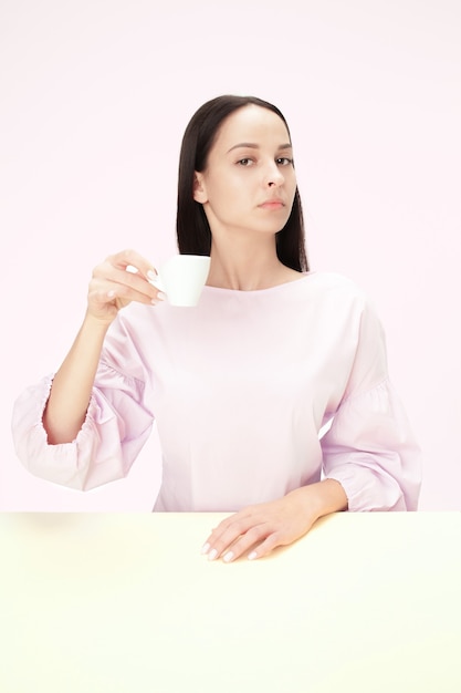 Bella donna sola che si siede allo studio rosa e che sembra triste tenendo in mano la tazza di caffè.