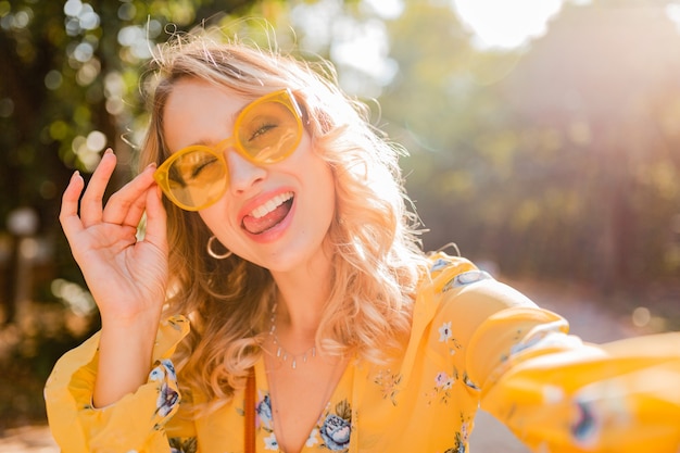 Bella donna sorridente alla moda bionda con l'espressione del fronte divertente in camicetta gialla che indossa occhiali da sole che fanno la foto del selfie