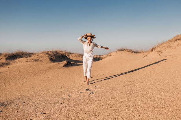 Bella donna sorridente alla moda che funziona nella sabbia del deserto in vestito bianco che porta il cappello di paglia sul tramonto