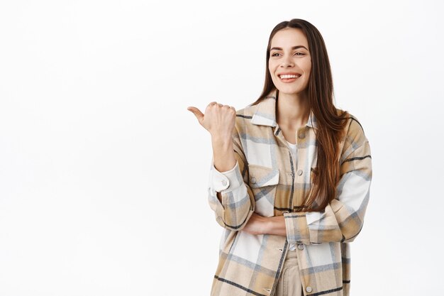 Bella donna sorridente che indica e guarda da parte, mostrando il logo, fissa compiaciuta lo spazio della copia con l'offerta promozionale del prodotto, in piedi contro il muro bianco