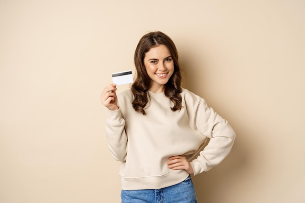 Bella donna sorridente che mostra carta di credito, concetto di shopping, pagamento contactless, in piedi su sfondo beige.