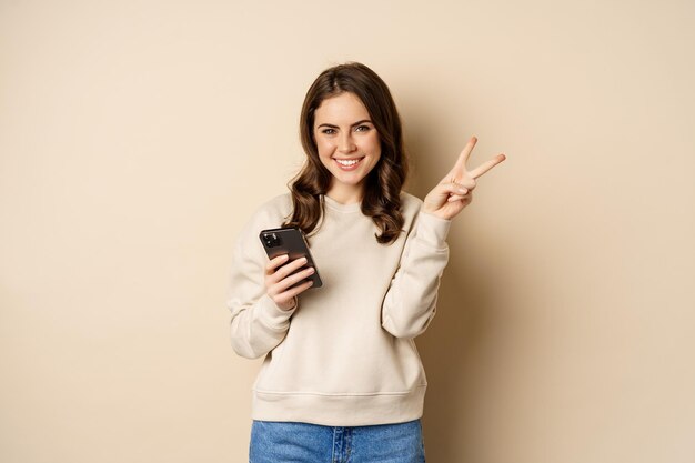 Bella donna sorridente che mostra il segno di pace, tenendo il telefono cellulare, in piedi su sfondo beige. Copia spazio