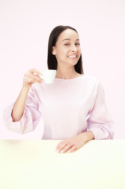 Bella donna sorridente che si siede allo studio rosa e che sembra felice che tiene la tazza di caffè in mano. Closeup ritratto tonico in stile minimalista