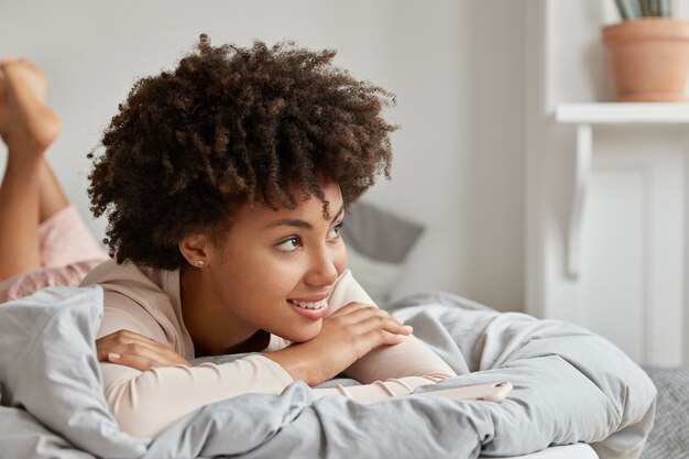 Bella donna sorridente con la pelle scura, ha un'espressione gioiosa, uno sguardo sognante, giace a pancia in giù a letto, gode di un buon riposo a casa, pianifica qualcosa in mente per i prossimi fine settimana, si sente rilassata. Ora di andare a letto