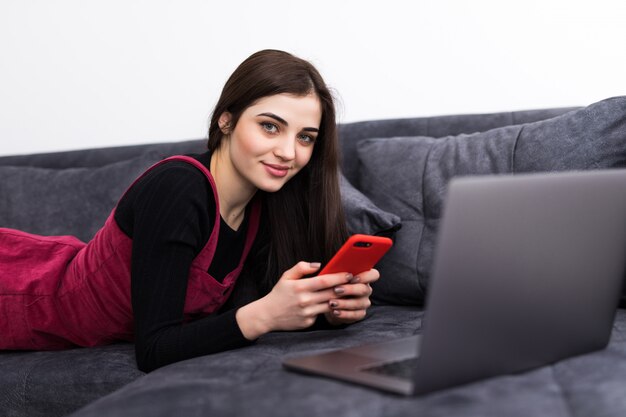 Bella donna sorridente dei giovani che si siede sul telefono e sul computer portatile di uso del sofà