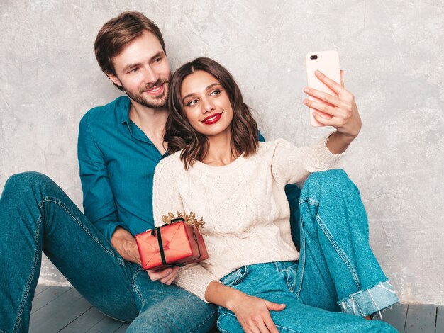 Bella donna sorridente e il suo bel ragazzo. Famiglia allegra felice che posa vicino alla parete grigia. San Valentino. Modelli che abbracciano e regalano alla sua ragazza una confezione regalo.