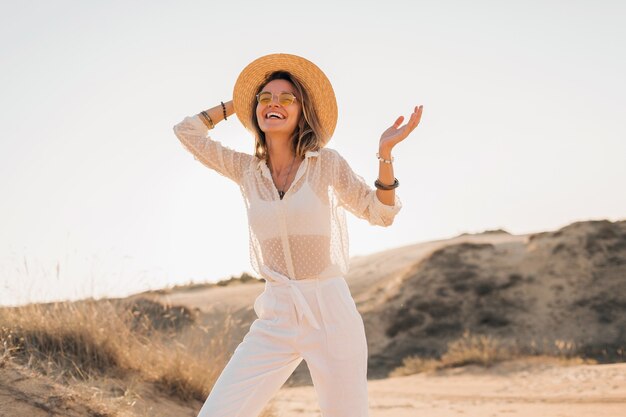 Bella donna sorridente felice alla moda che posa nella sabbia del deserto in vestito bianco che porta cappello di paglia e occhiali da sole sul tramonto