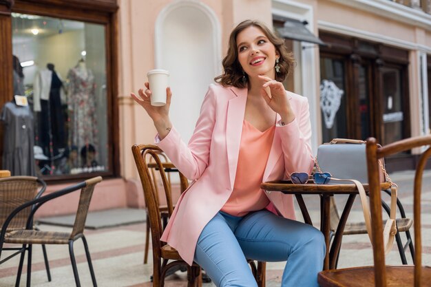 Bella donna sorridente in abito elegante seduto a tavola indossando giacca rosa, romantico stato d'animo felice, data in caffetteria, tendenza moda primavera estate, bere caffè, fashionista