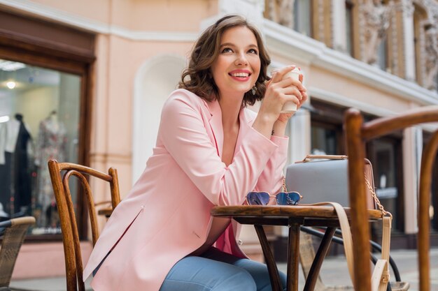 Bella donna sorridente in abito elegante seduto al tavolo che indossa giacca rosa, romantico stato d'animo felice, aspettando il fidanzato a un appuntamento al bar, tendenza moda primavera estate, bere caffè