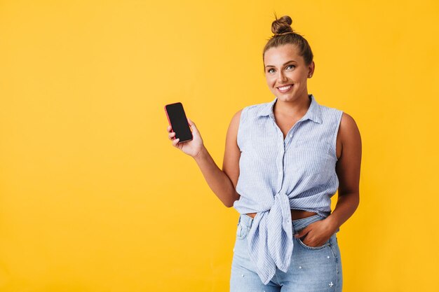 Bella donna sorridente in camicia che guarda felicemente a porte chiuse mentre mostra il nuovo cellulare su sfondo giallo