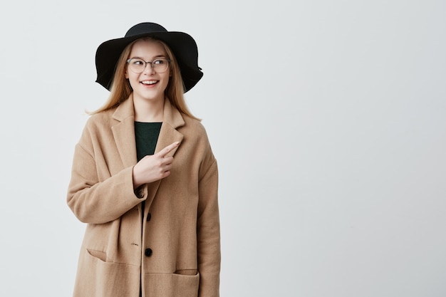 Bella donna sorridente in cappotto sopra maglione verde e occhiali che punta al muro bianco bianco mentre dimostrando qualcosa. Giovane femmina allegra che indica con il dito anteriore su fondo grigio.