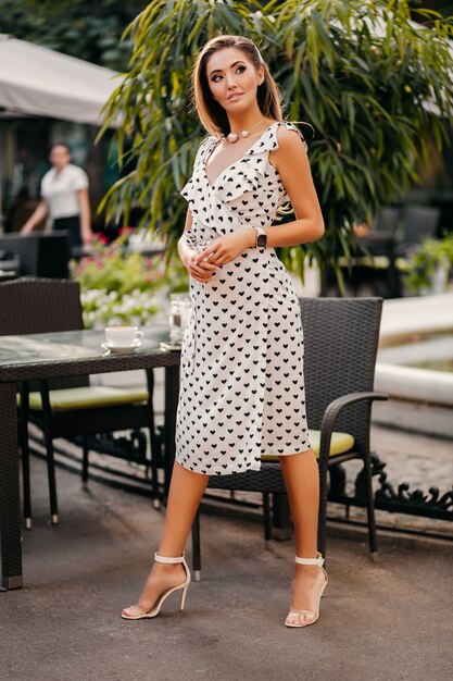 Bella donna sorridente vestita in abito stampato bianco in stile romantico in posa in street cafe il giorno pieno di sole