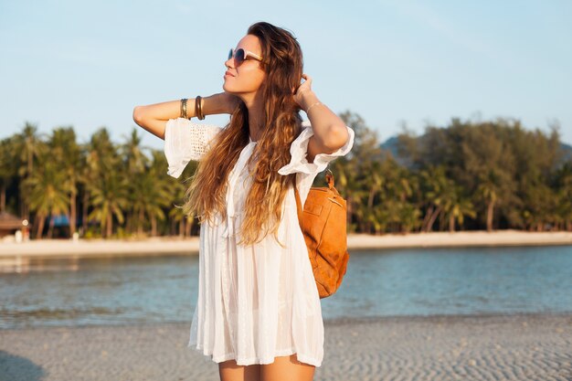 Bella donna sottile in vestito di cotone bianco che cammina sulla spiaggia tropicale sul tramonto che tiene lo zaino in pelle.