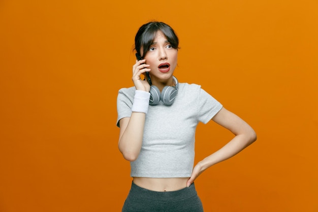 Bella donna sportiva in abbigliamento sportivo con le cuffie che parlano al telefono cellulare che sembra confusa in piedi su sfondo arancione