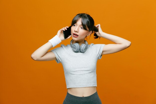 Bella donna sportiva in abbigliamento sportivo con le cuffie che parlano al telefono cellulare che sembra sicura e incuriosita in piedi su sfondo arancione