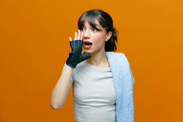 Bella donna sportiva in abbigliamento sportivo con un asciugamano sulla spalla in guanti tiene la mano vicino alla bocca come chiamare qualcuno in piedi su sfondo arancione